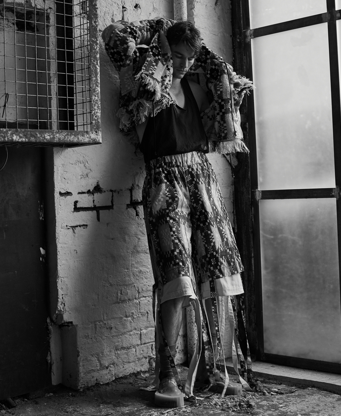 Black and white portrait of a young adult posing in a patterned jacket and shorts near a window in an aged interior with brick walls.