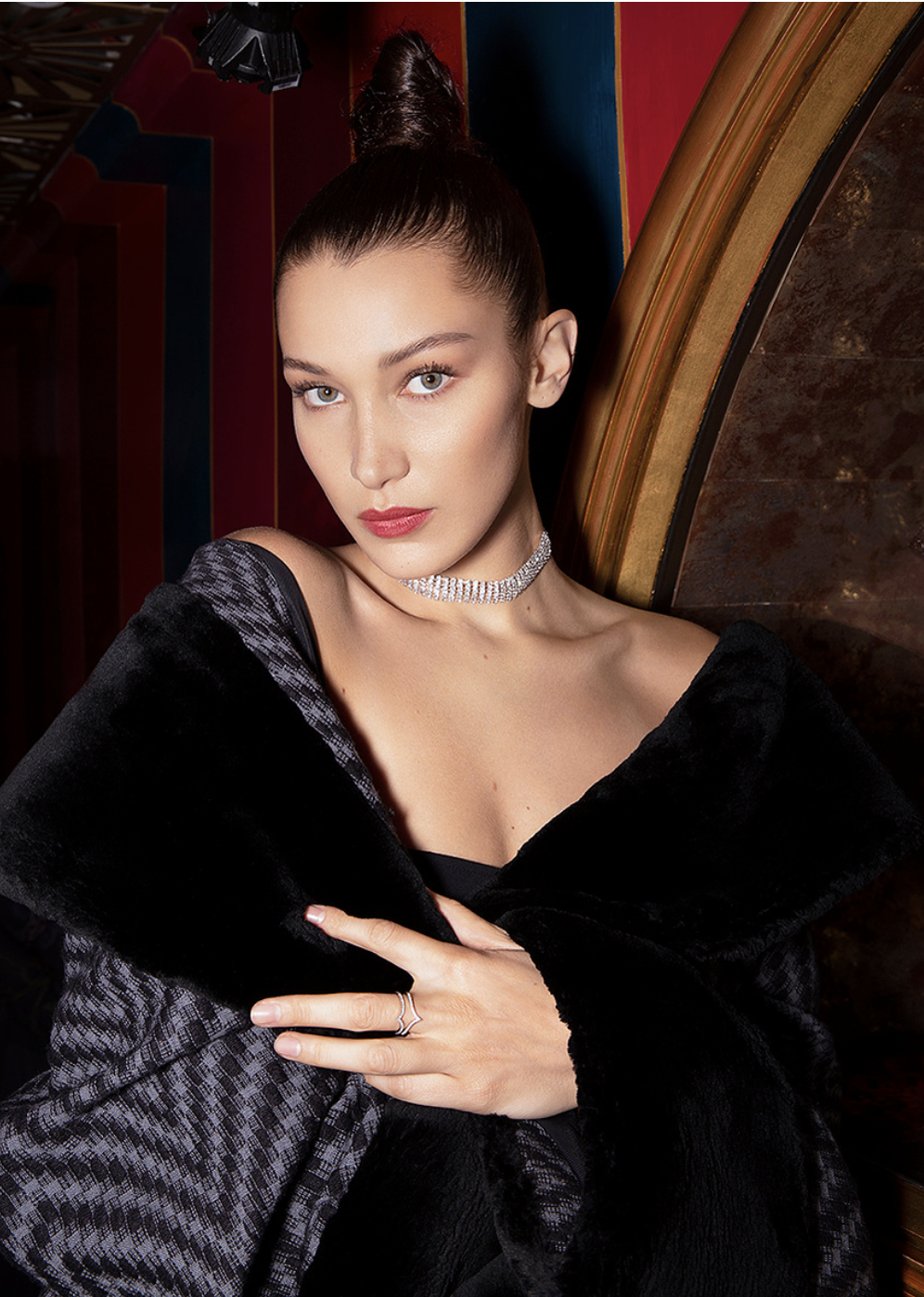 Portrait of an adult woman with a high bun wearing a black and gray faux fur shawl and diamond choker indoors with striped background.