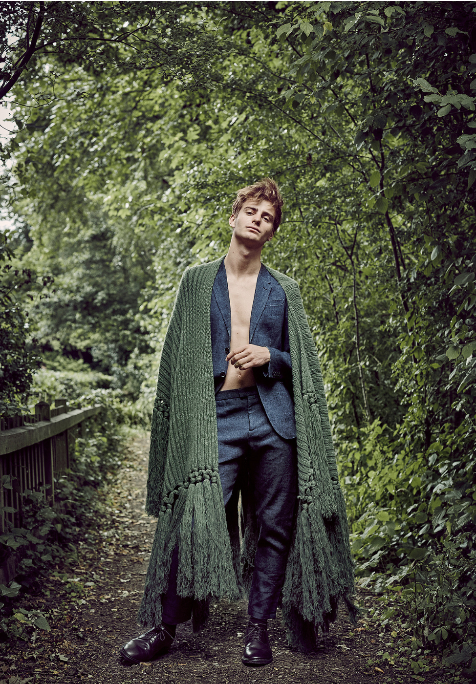 Young adult man poses on a dirt path in an open suit with a knitted green shawl in a wooded outdoor environment.