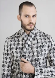 Man with a buzz cut and beard wearing a black and white geometric patterned coat against a neutral studio background.