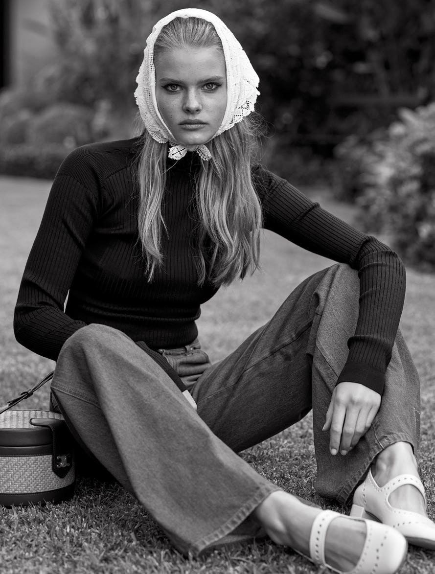 Monochrome portrait of a woman in a headscarf, dark top, jeans, and Mary Jane shoes, sitting on grass with a round woven bag.
