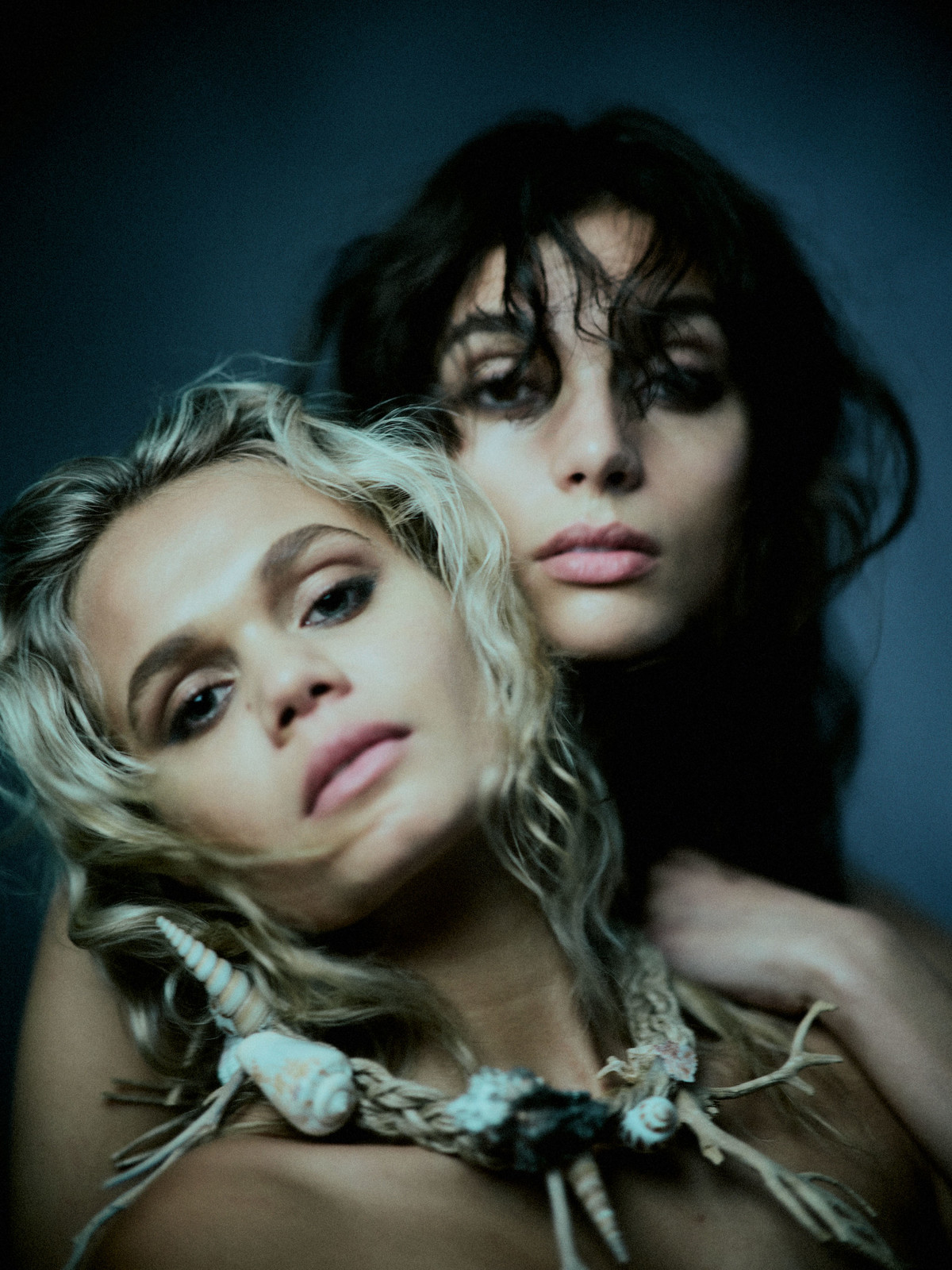 Two women pose in a close-up portrait with one wearing a seashell necklace in a moody and dimly lit indoor setting with dark background.