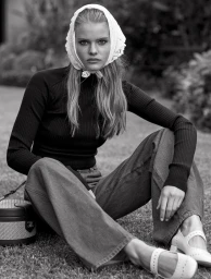 Black and white portrait of a blonde woman wearing a lace headscarf, dark turtleneck sweater, and wide-legged pants sitting outdoors on grass.