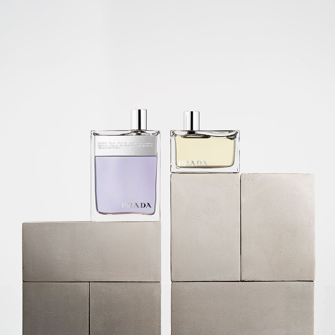Two Prada fragrances in clear glass bottles are displayed on gray concrete blocks against a white backdrop in a minimalist composition.