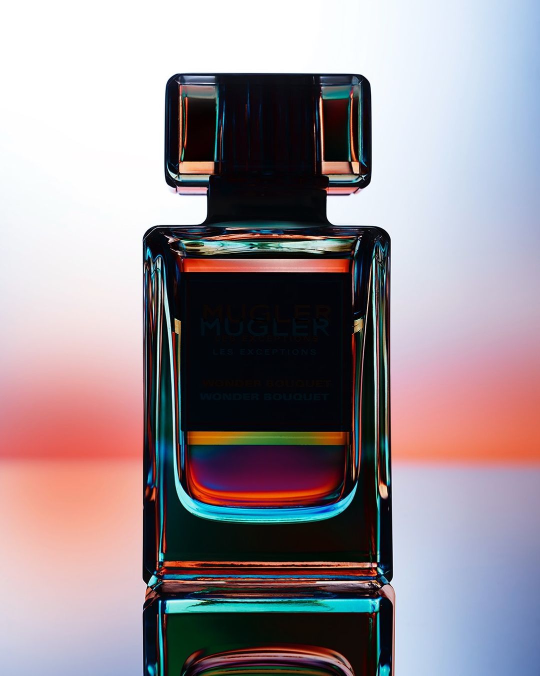 Close-up studio shot of a glass MUGLER fragrance bottle with a black cap, a colorful light band, and vibrant reflections on a glossy surface.