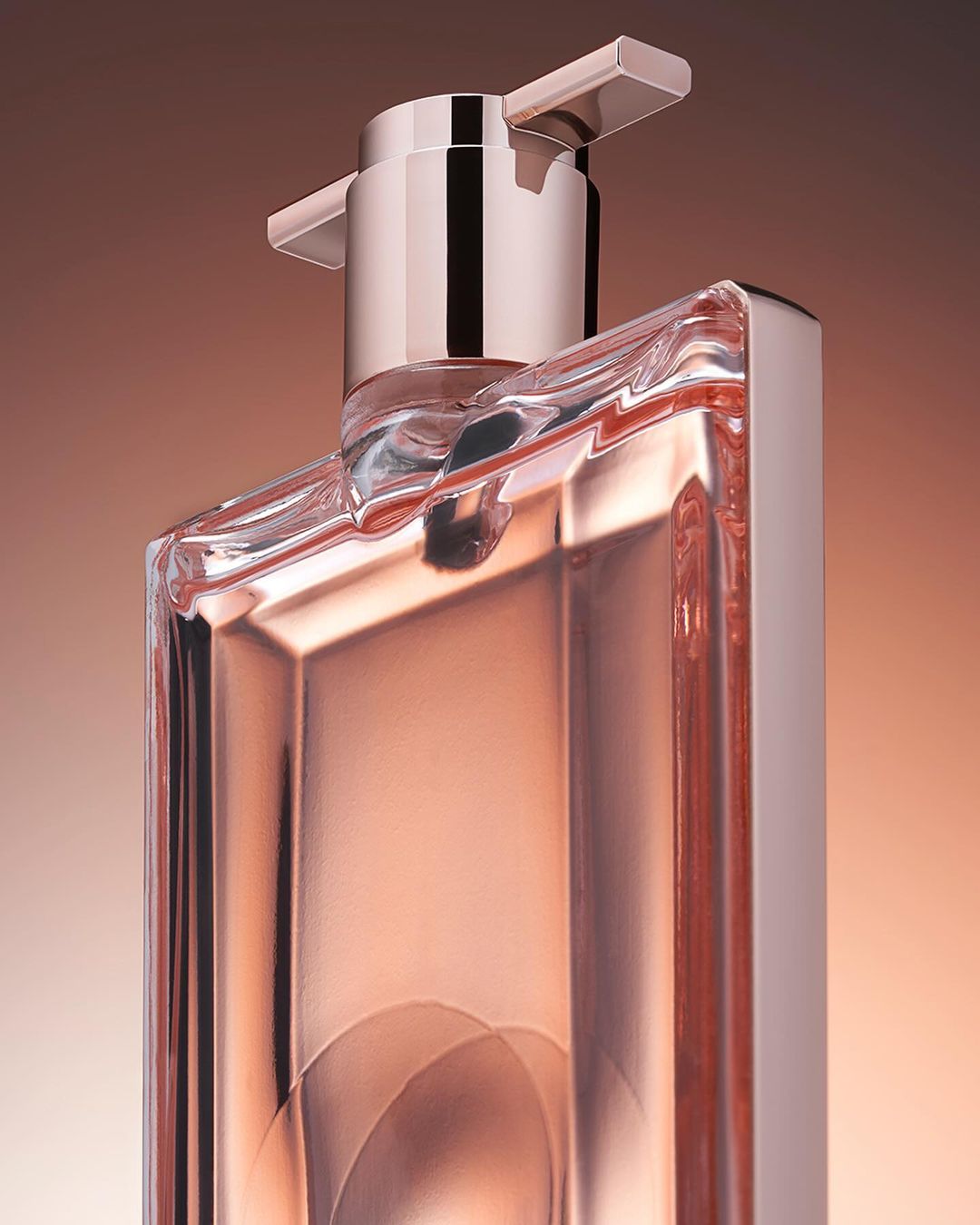 Close-up studio shot of a clear glass perfume bottle with rose gold accents against a gradient beige and brown background.