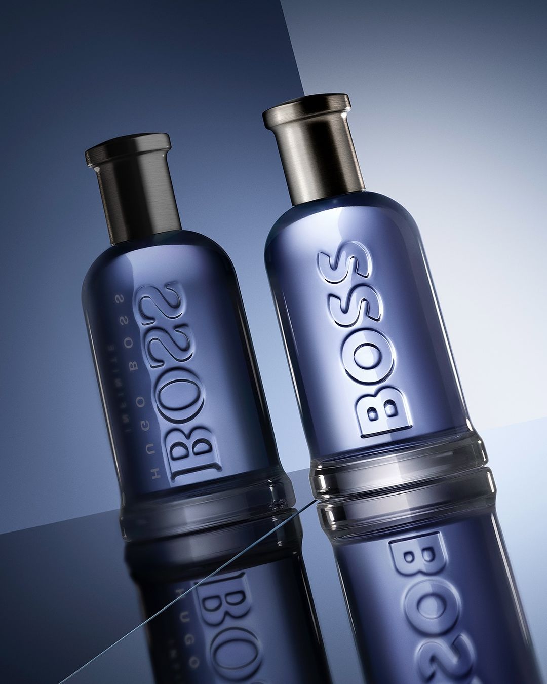 Two blue Hugo Boss Bottled Parfum bottles on a reflective surface against a gradient blue and white background, studio lighting.