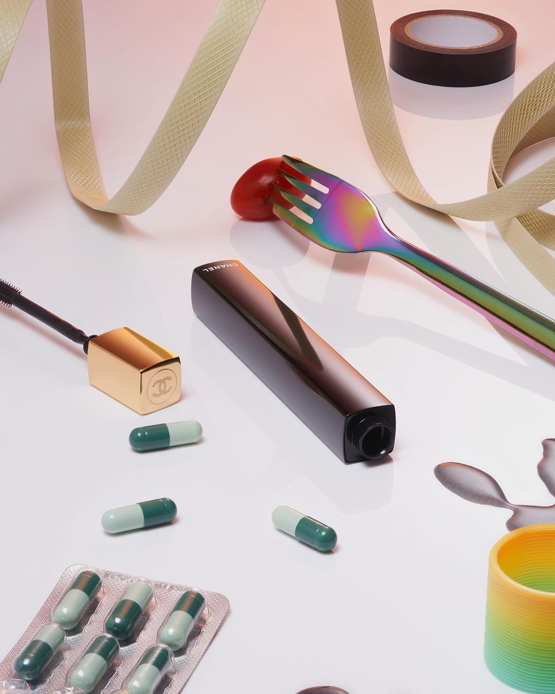 Still life studio shot featuring makeup products, capsules, a rainbow fork with a tomato, and a beige ribbon on a white background.