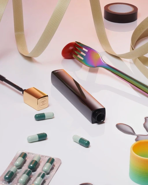 Still life studio shot featuring makeup products, capsules, a rainbow fork with a tomato, and a beige ribbon on a white background.