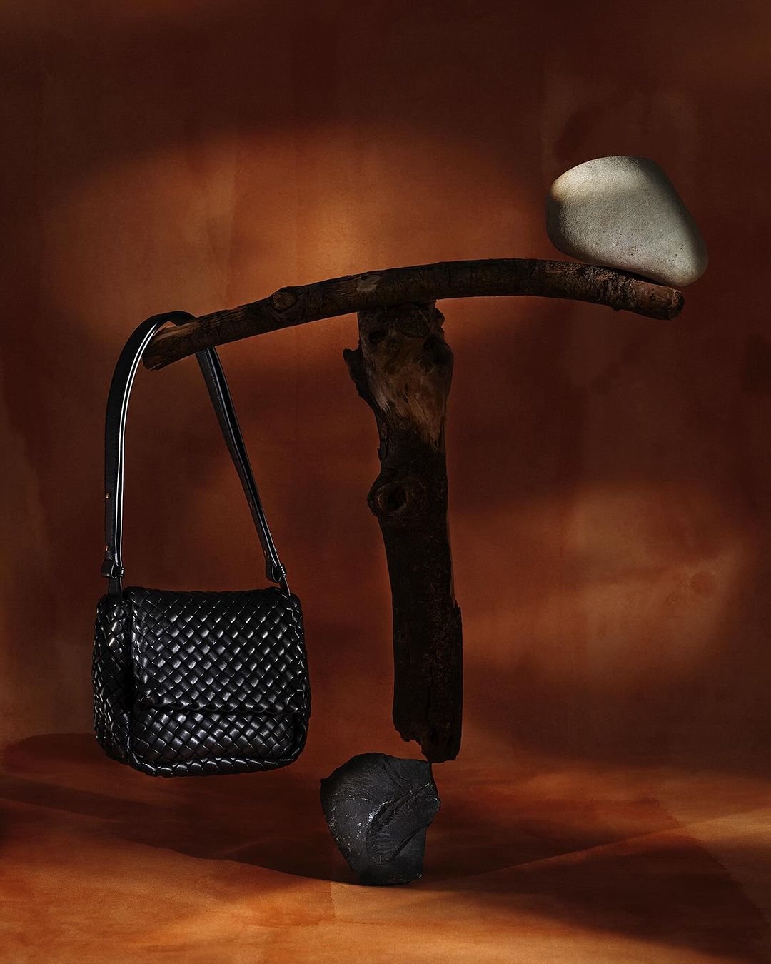 Studio shot of a black woven handbag balanced by rocks and a wooden branch against a tan-brown gradient background. Fashion still life.