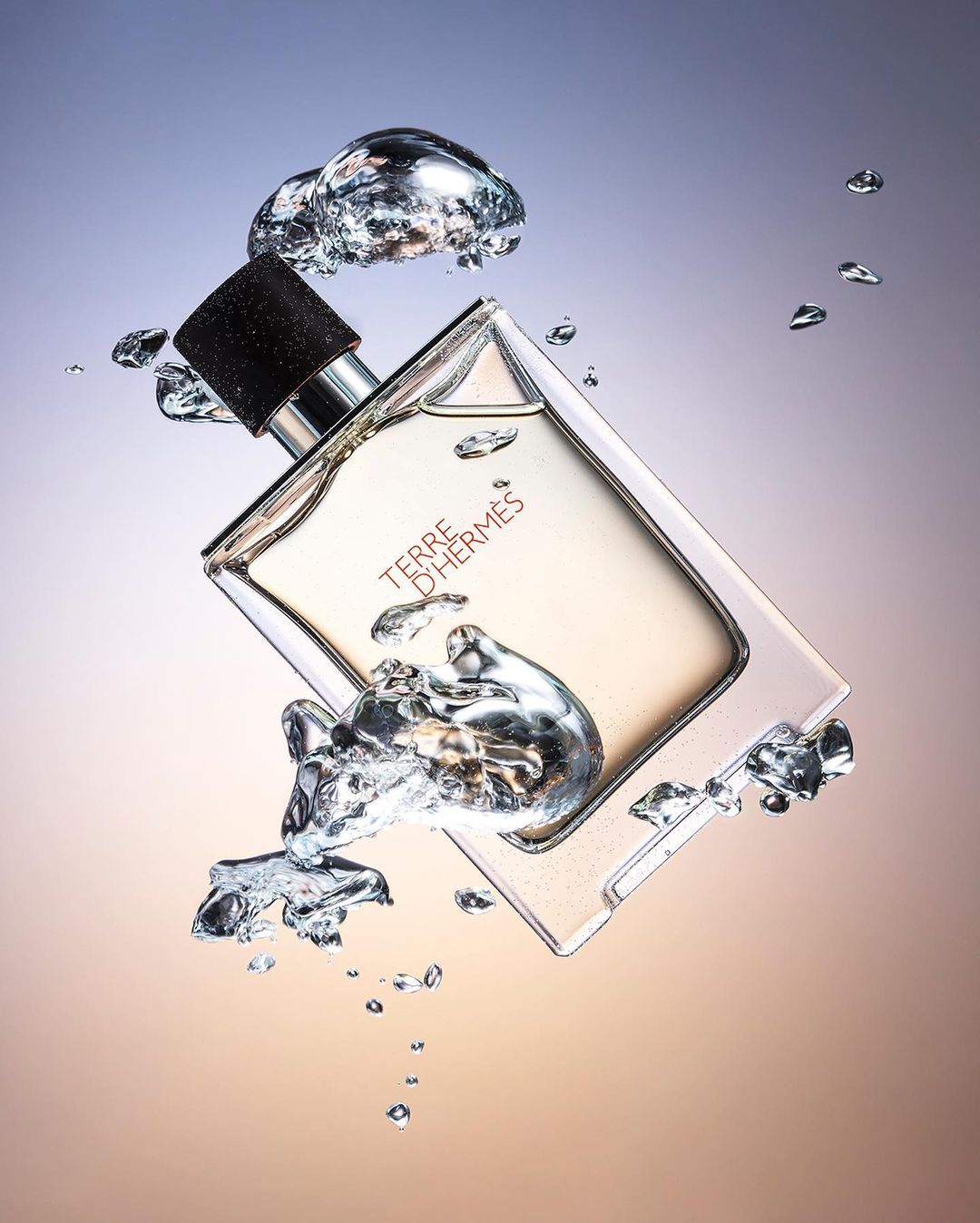 A fragrance bottle with a black cap is shown in studio lighting, surrounded by water droplets. The product is Terre d'Hermès.