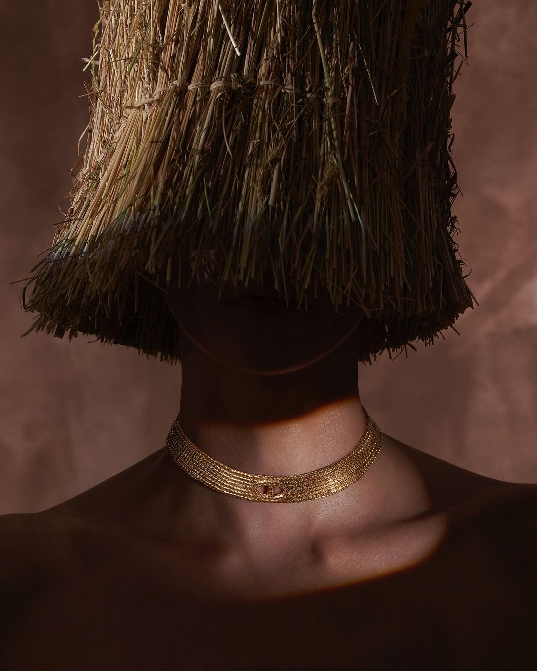 Close-up of a person wearing a golden choker necklace and a hat made of hay against a light brown background.