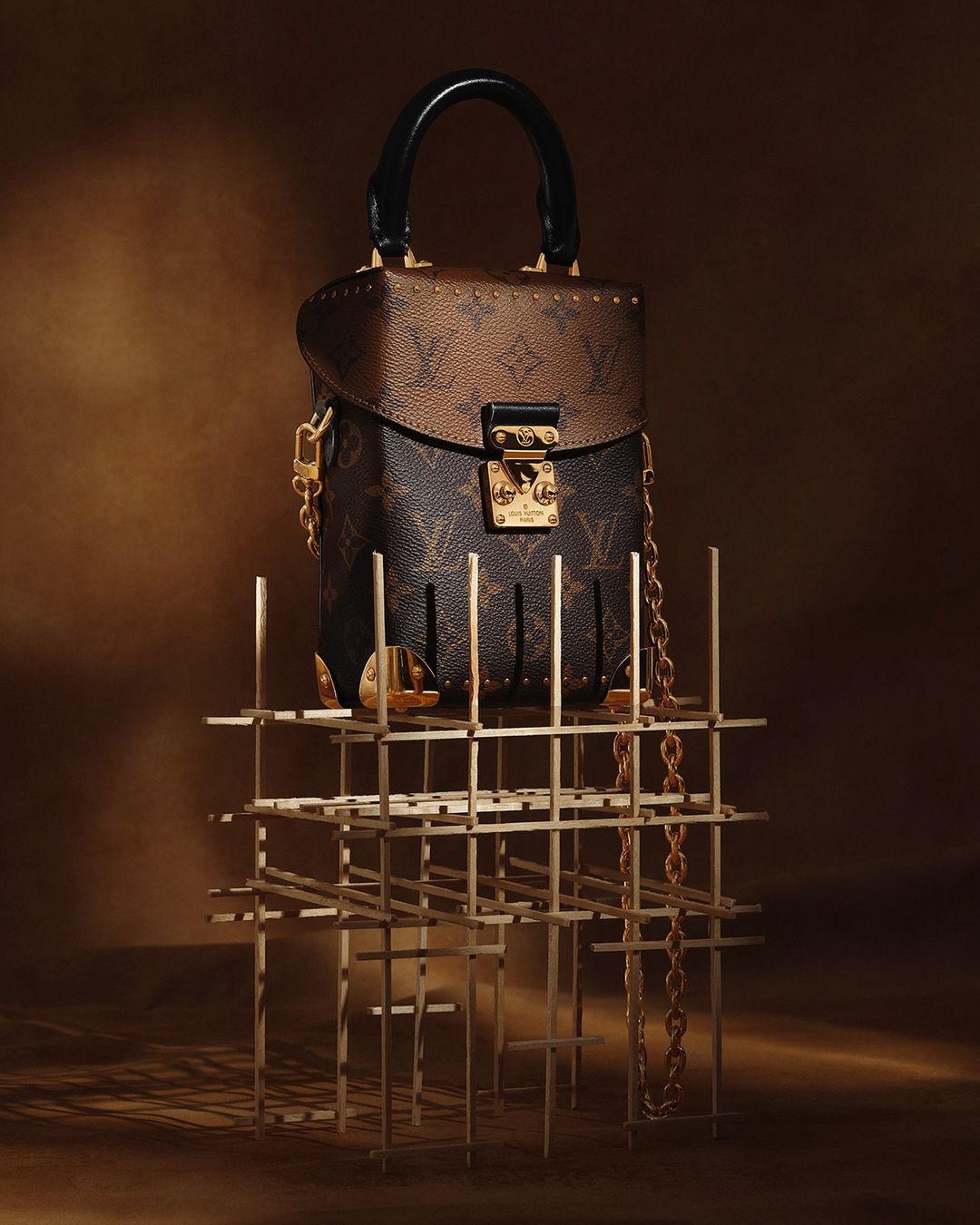 Luxury Louis Vuitton top-handle bag with chain strap, displayed on a gold metal geometric stand against a neutral brown backdrop.