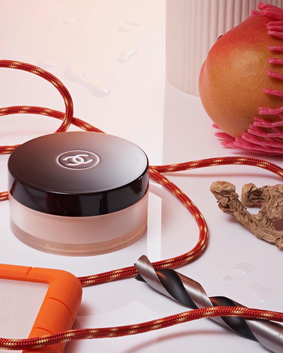 Chanel cream jar, drill bits, orange frame, ginger root and mango with pink netting on a reflective white surface.