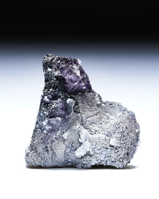 Close-up studio shot of a purple and gray mineral formation with white speckles, against a gradient gray and white background.