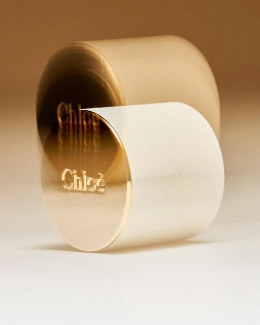 Close-up studio shot of a gold and white cylindrical container with 'Chloé' logo on a gradient neutral background, fashion-forward minimalist product.