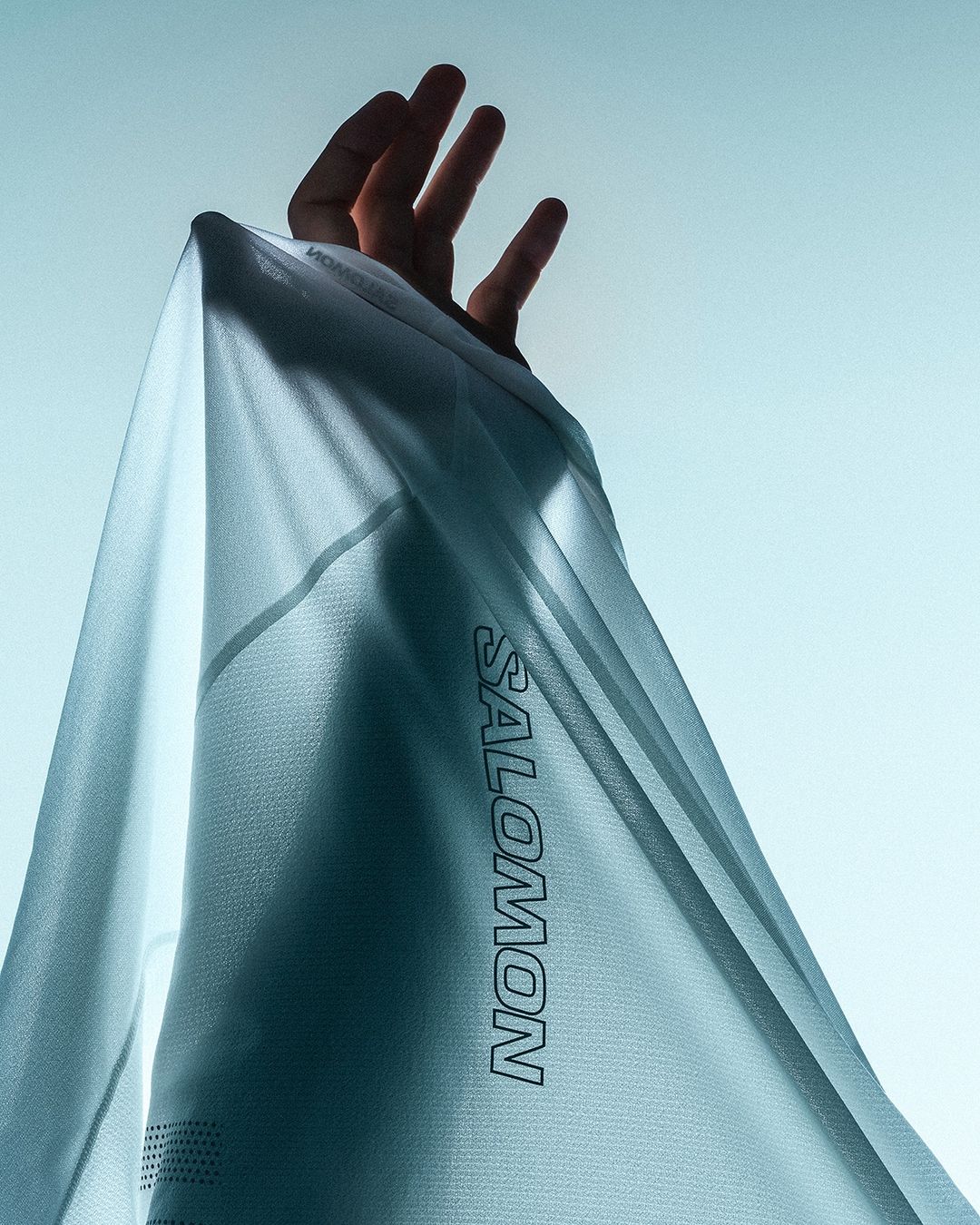 Close-up of a hand holding up a light blue Salomon brand athletic shirt against a bright, clear sky backdrop.
