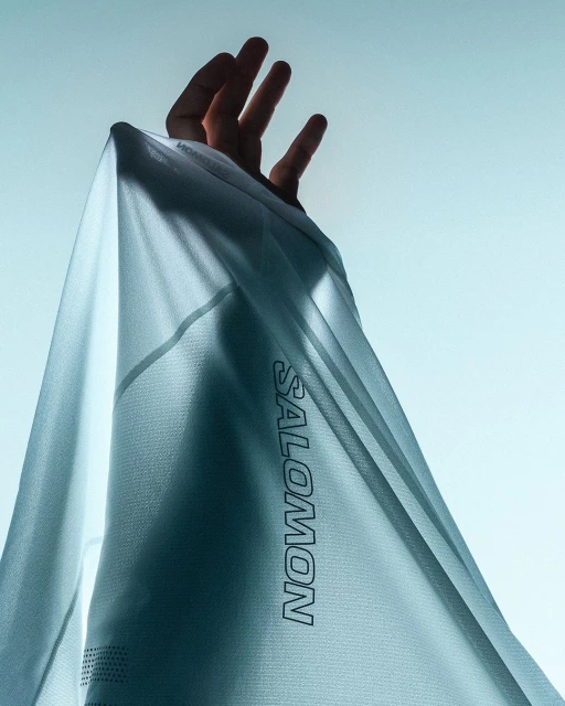Close-up of a hand holding up a light blue Salomon brand athletic shirt against a bright, clear sky backdrop.