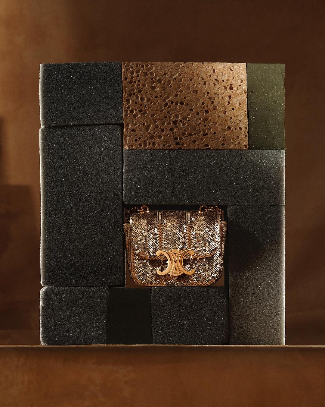 Studio shot of a gold sequined handbag with a gold metal logo, displayed within an arrangement of dark gray blocks.