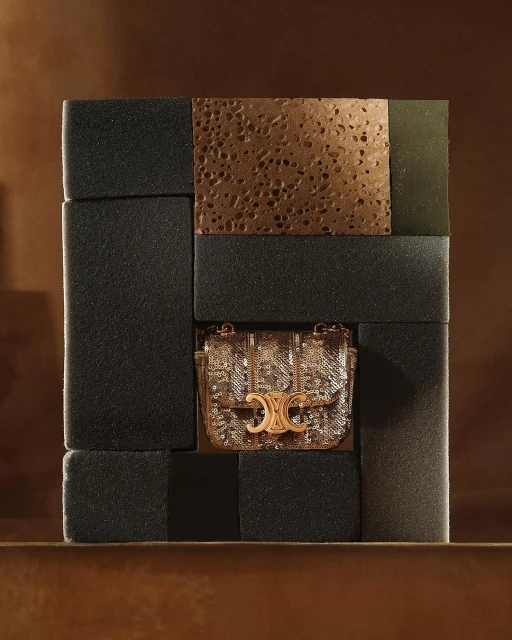Studio shot of a gold sequined handbag with a gold metal logo, displayed within an arrangement of dark gray blocks.