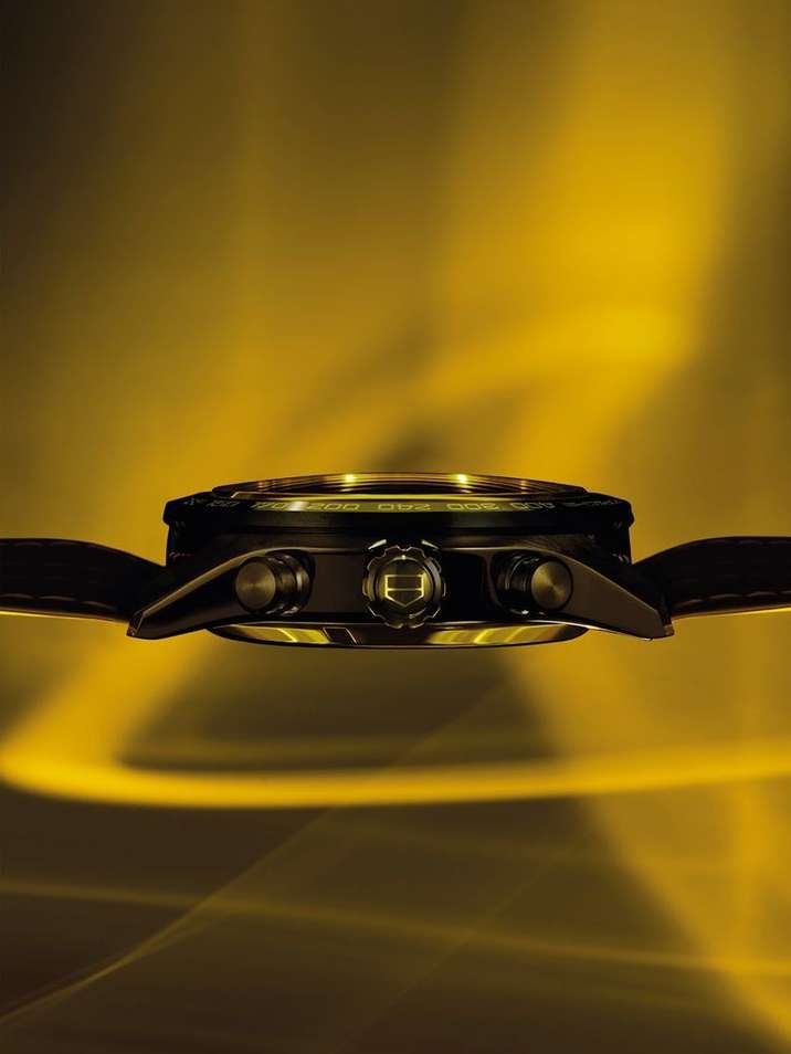 Side view of a black wristwatch with gold accents displayed against a blurred yellow background with wave-like lighting patterns.