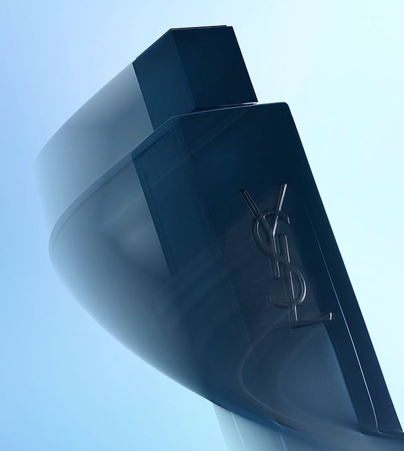 Close-up of a sleek, dark blue Yves Saint Laurent fragrance bottle with a metallic logo, against a soft blue gradient background.