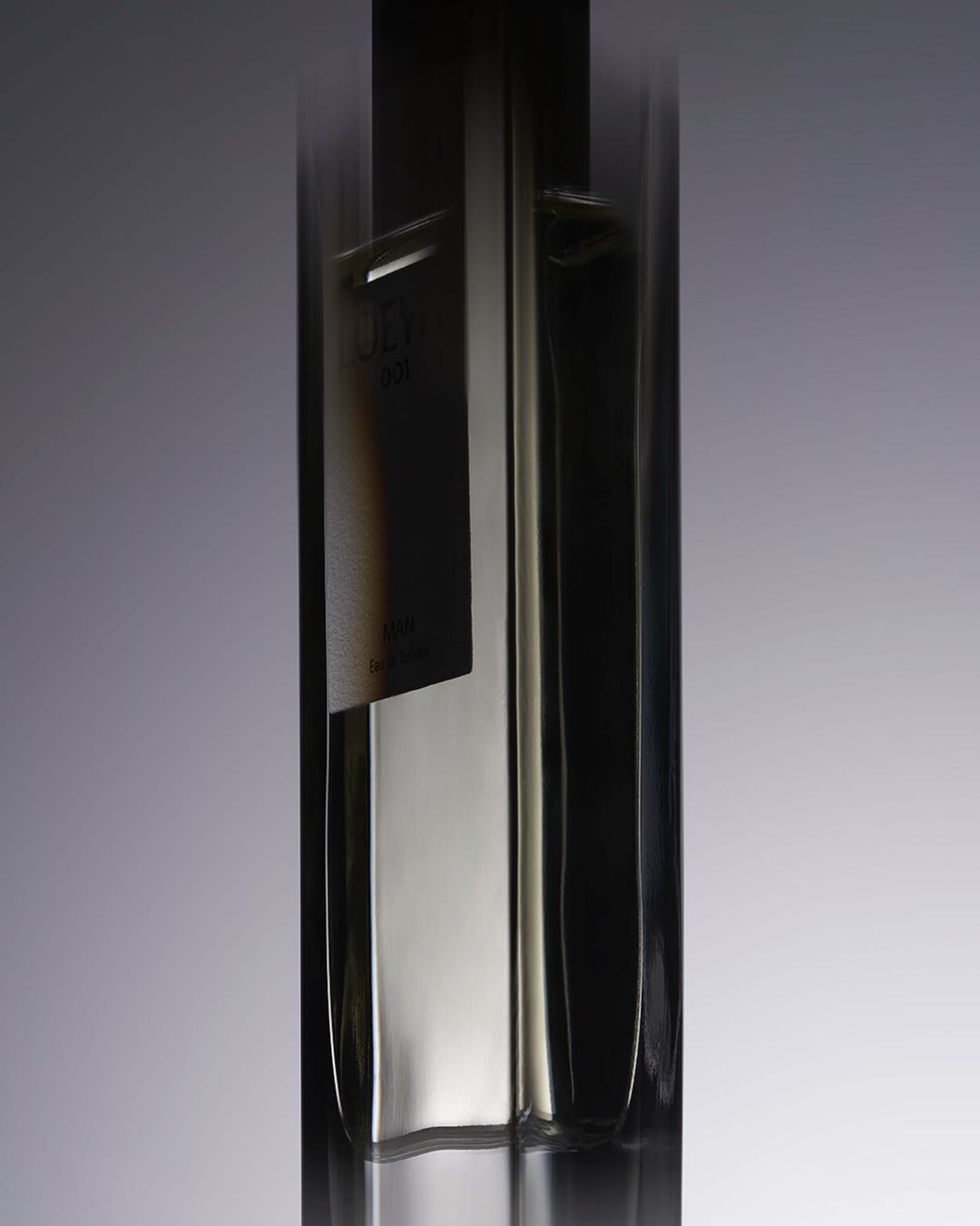 Close-up studio shot of a tall, clear glass bottle with a rectangular black label and reflective properties against a gradient background.