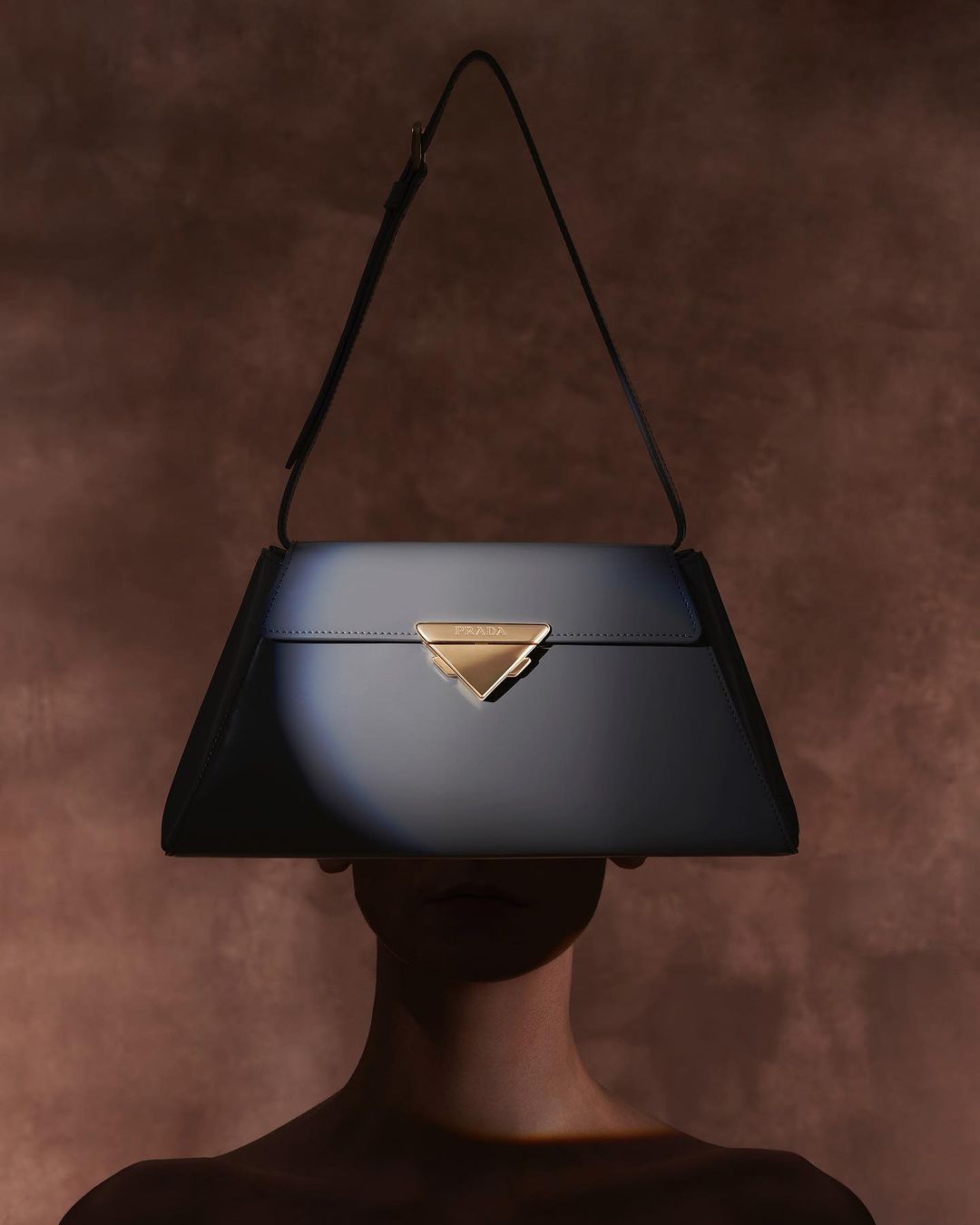 Fashion shot of a woman partially hidden behind a navy-blue trapezoid-shaped leather handbag with a triangular gold clasp.