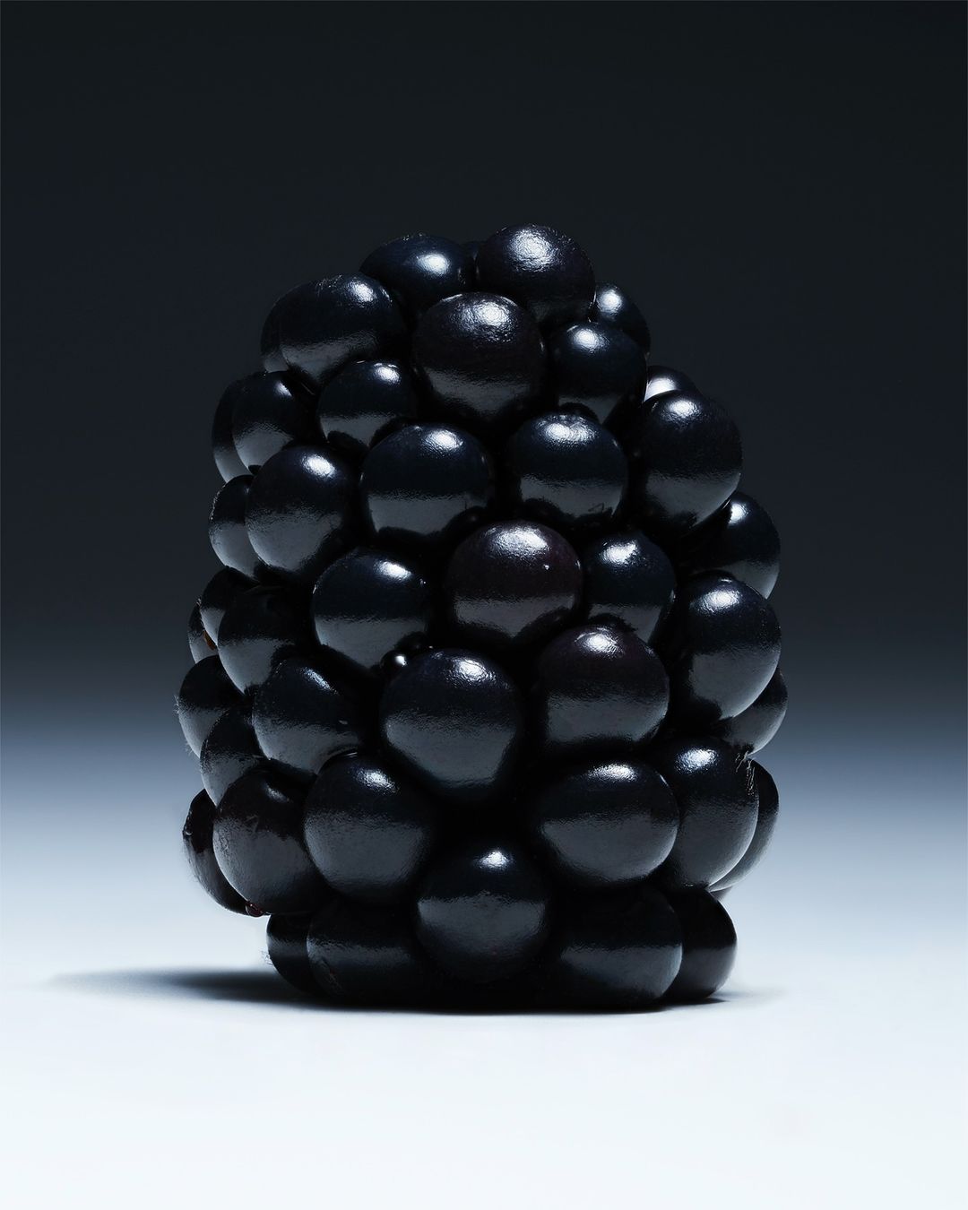 Close-up product photography showcases a single blackberry against a dark background, highlighting its dark purple color and textured surface.