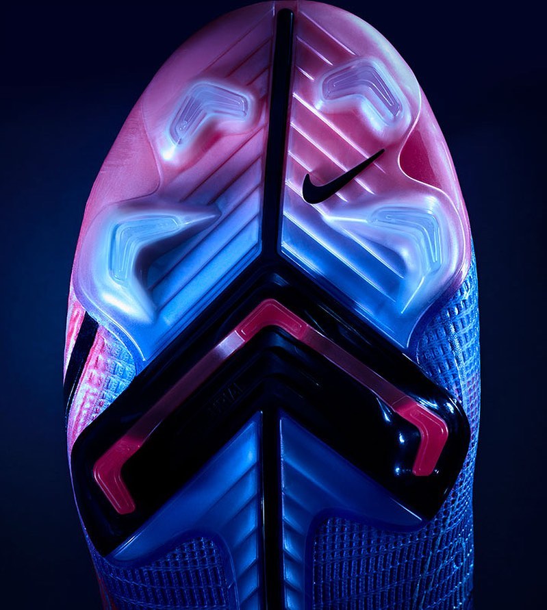 Close-up of the sole of a blue and pink Nike soccer cleat, showing the studs and Nike logo on a dark background.