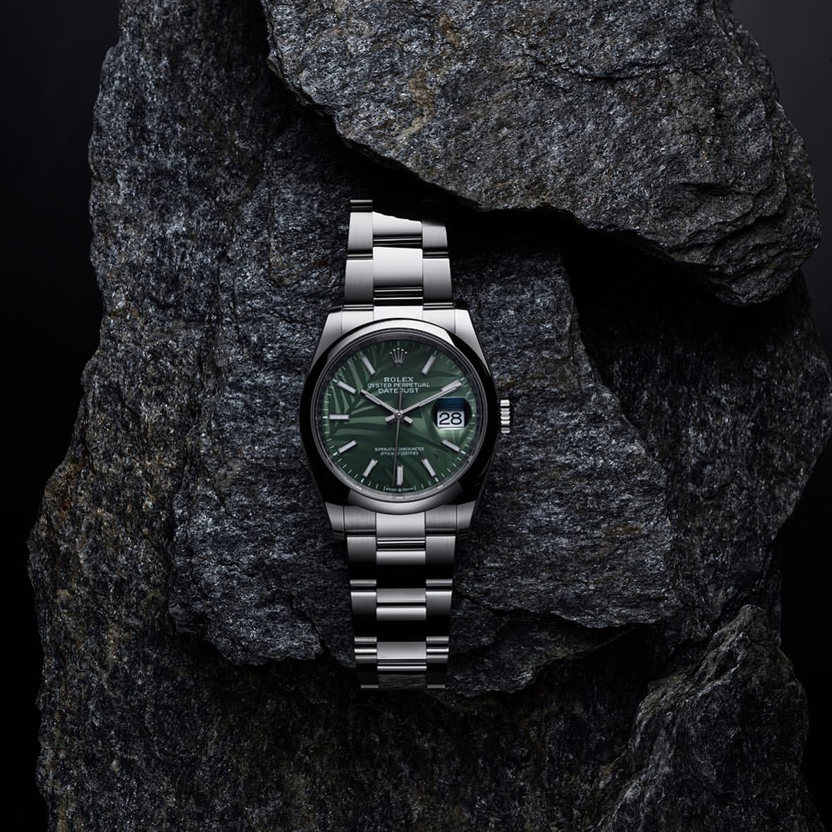 Silver Rolex Datejust watch with a green patterned dial rests on a textured grey rock against a dark background in a studio product shot.