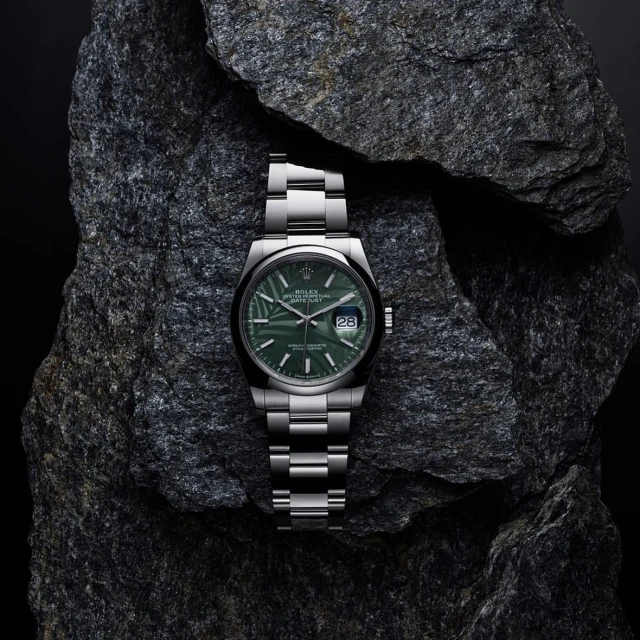 Silver Rolex Datejust watch with a green patterned dial rests on a textured grey rock against a dark background in a studio product shot.