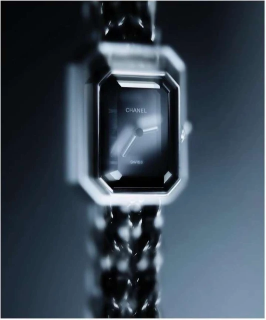 Close-up of a square, black Chanel wristwatch with a silver chain band on a gradient grey background, luxury fashion accessory.