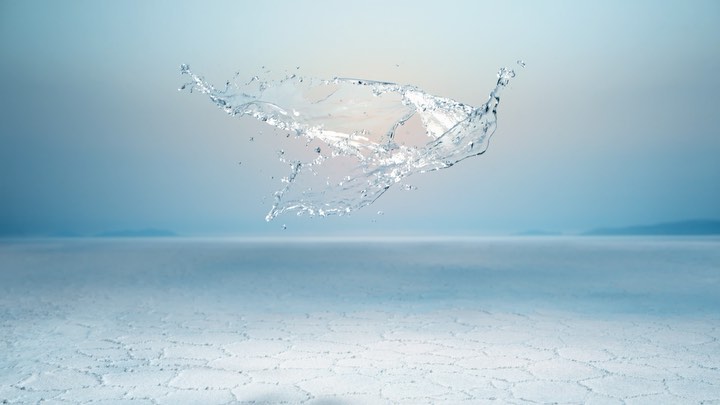 Water splash suspended in mid-air, blue sky background with horizon over cracked, pale desert surface in horizontal landscape.
