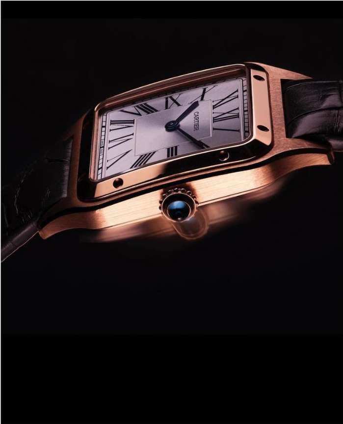 Close-up of a rose gold Cartier watch with a white face and black leather strap set against a dark, blurred background.
