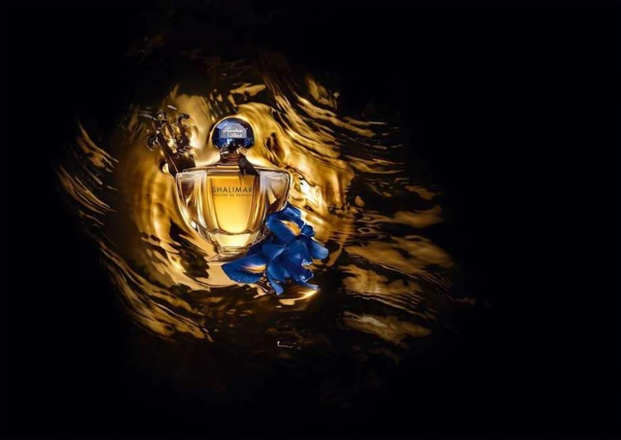 A glass Guerlain Shalimar perfume bottle sits with blue flowers in the foreground and a background of bronze and black swirls.