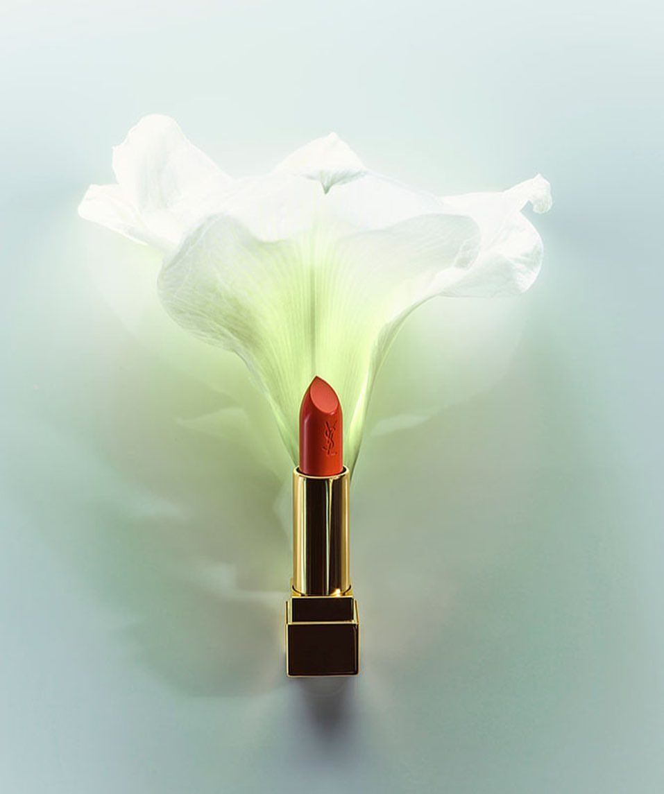 A close-up studio shot of vibrant orange lipstick with gold casing, cradled by the delicate white petals of a luminous calla lily flower.