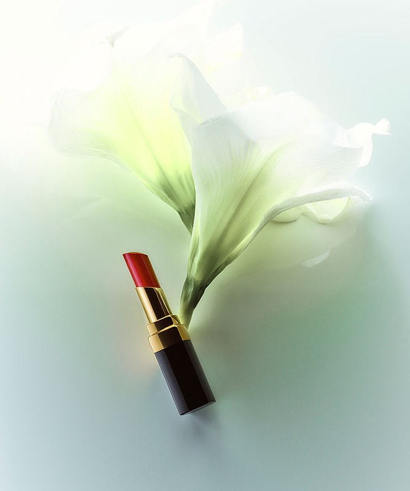 Close-up shot of a red lipstick tube positioned against a backdrop of elegant white lily flowers on a soft, light-toned surface.