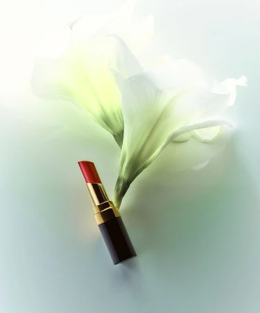 Close-up shot of a red lipstick tube positioned against a backdrop of elegant white lily flowers on a soft, light-toned surface.