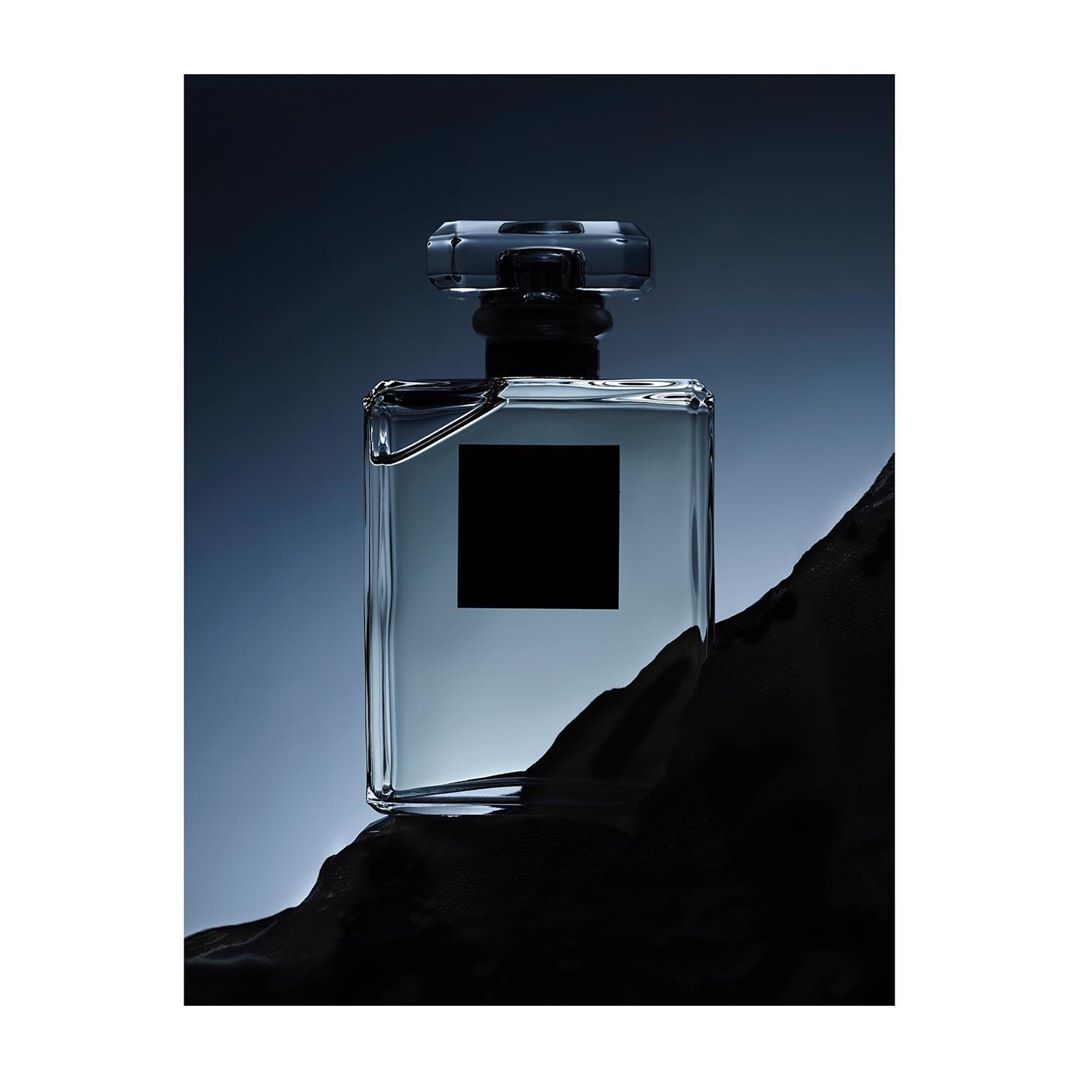 Product shot of a translucent perfume bottle with a dark cap, set against a dim, gradating blue background with a rough, dark stone base.