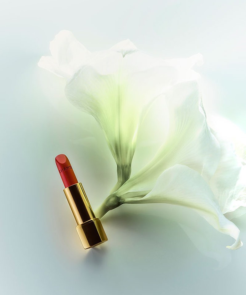 A red and gold lipstick tube lies next to a white flower against a gradient pale-blue background in a beauty or fashion still-life photo.