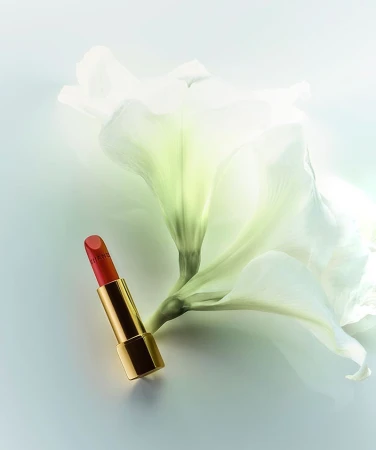 A red and gold lipstick tube lies next to a white flower against a gradient pale-blue background in a beauty or fashion still-life photo.