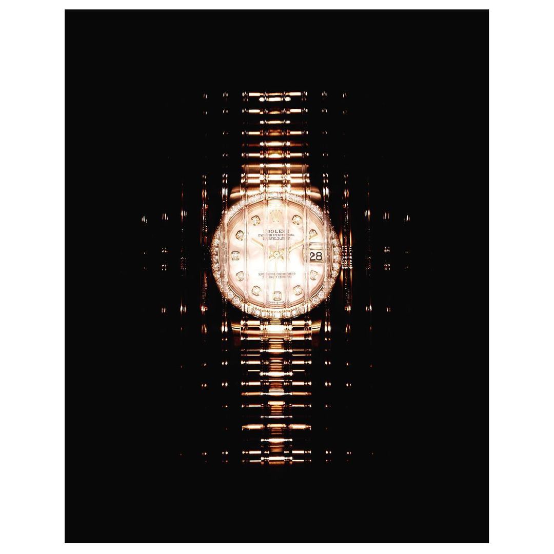 Luxury gold wristwatch with a gold band on a dark background with a radial pattern emanating from the face of the watch.