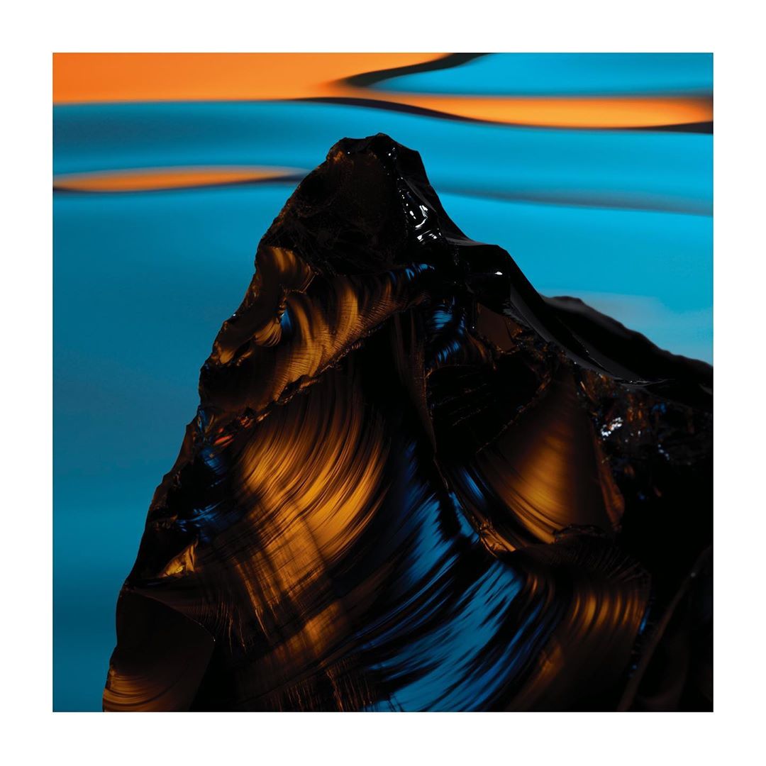 Dramatic studio shot of a large, dark, polished obsidian rock formation with striking blue and orange lighting creating a surreal effect.