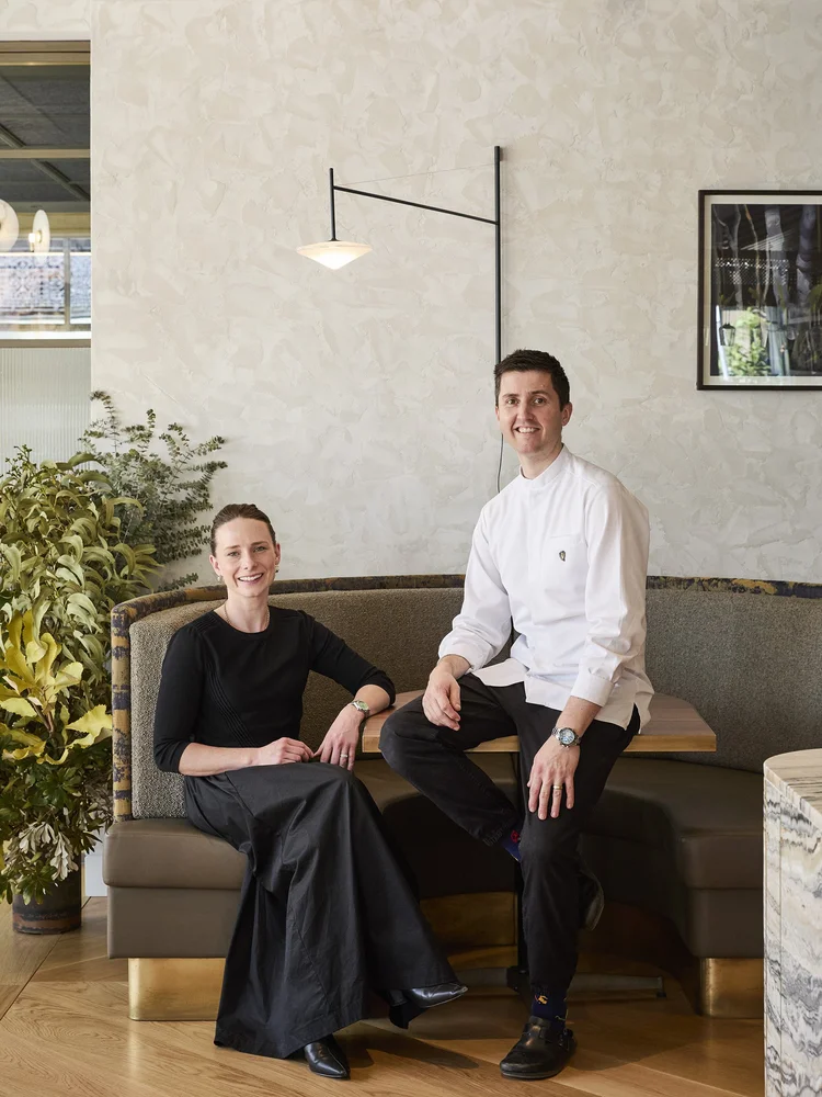 Indoor portrait of two adults posing in a restaurant: an adult female seated in a black top and long skirt and an adult male wearing a chef's coat.