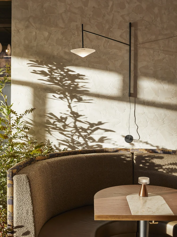 Indoor restaurant setting featuring a lit wall lamp, a patterned booth, a wood table with a small lamp, and light from tree shadows.
