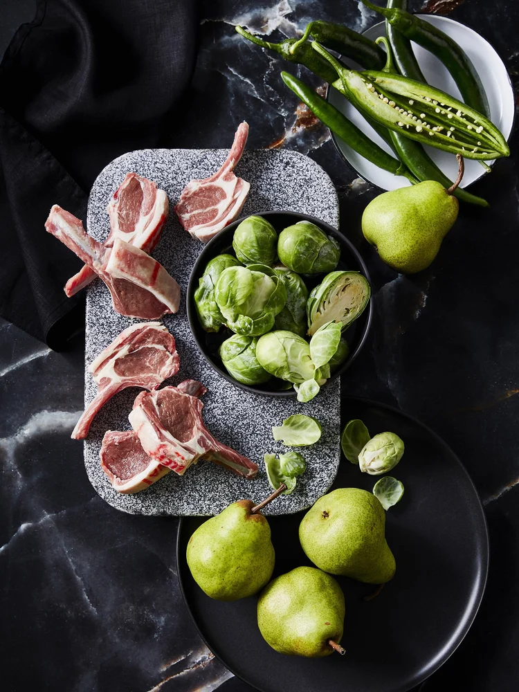 Overhead view of lamb chops, green pears, Brussels sprouts, and green chili peppers arranged on a grey stone slab and black surface.