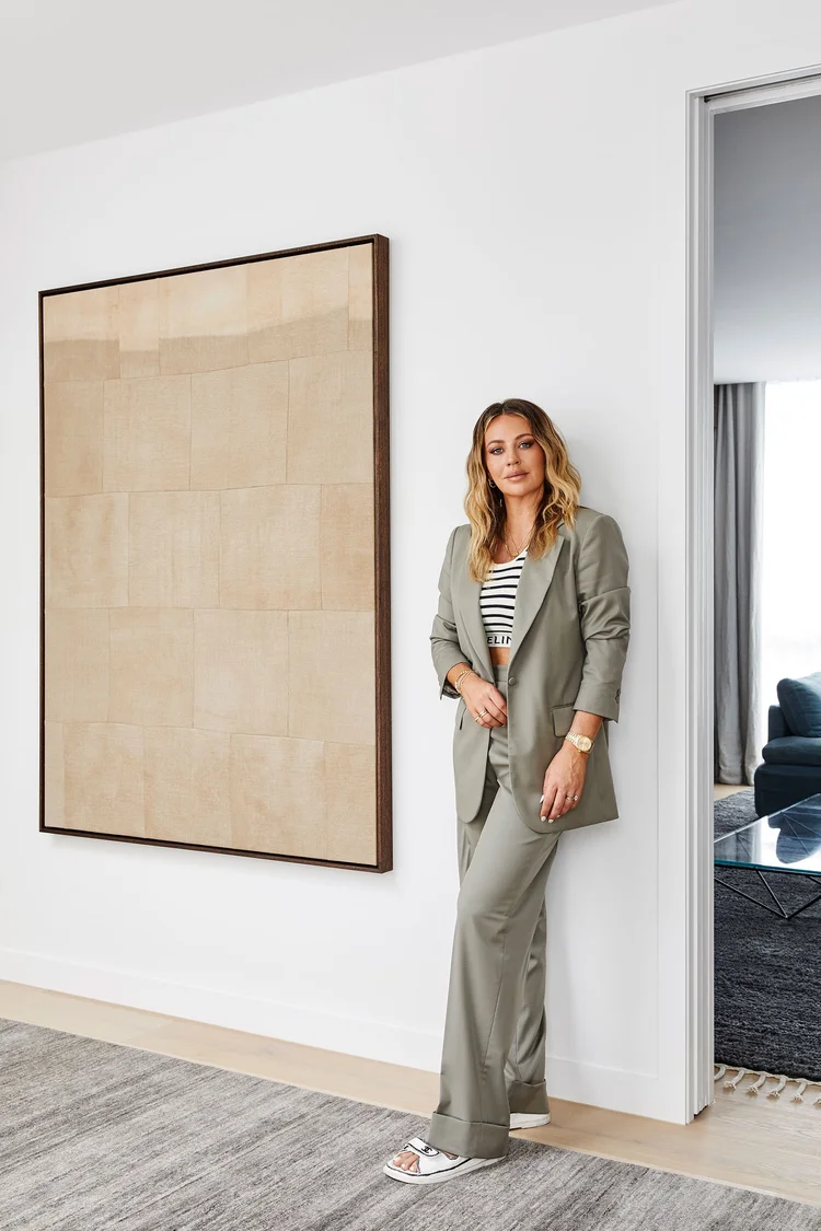Blonde woman in an olive suit leans against a white wall next to an abstract painting in an interior space with a gray rug.