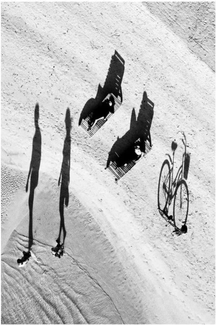 Aerial shot of beach with silhouettes of people, chairs, and a bicycle creating a black-and-white pattern in the sand.