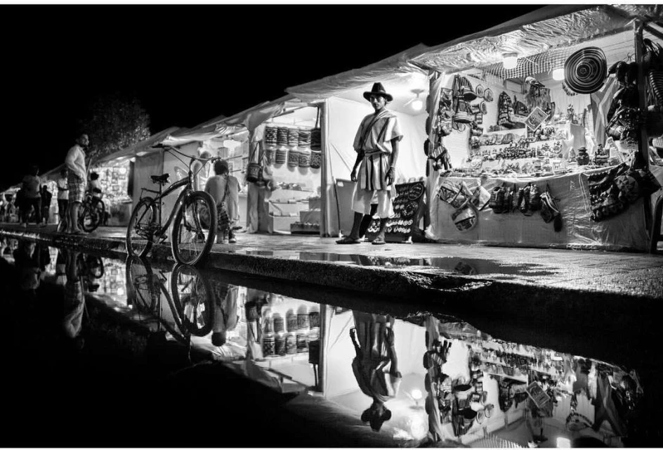 Black and white shot of a man in a hat standing next to a booth at an outdoor market at night, with a bicycle and reflections.